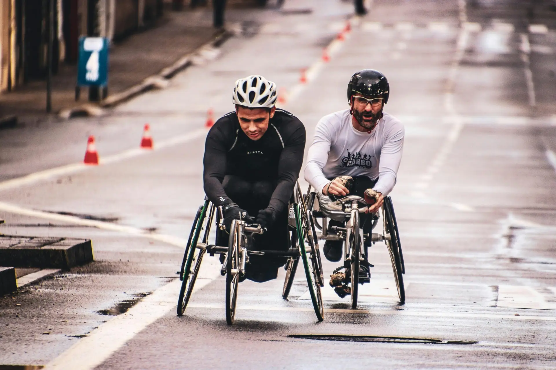 Two men compete in a wheelchair race on a wet urban street, showcasing endurance and teamwork.