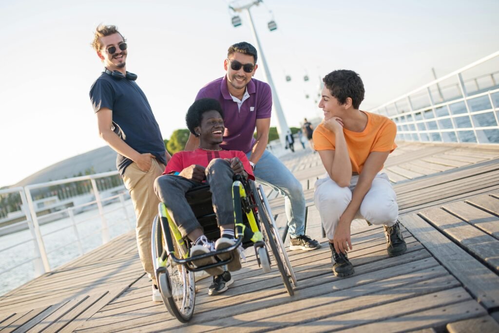 Four friends, including a man in a wheelchair, laughing and enjoying a sunny day outdoors on a wooden deck.